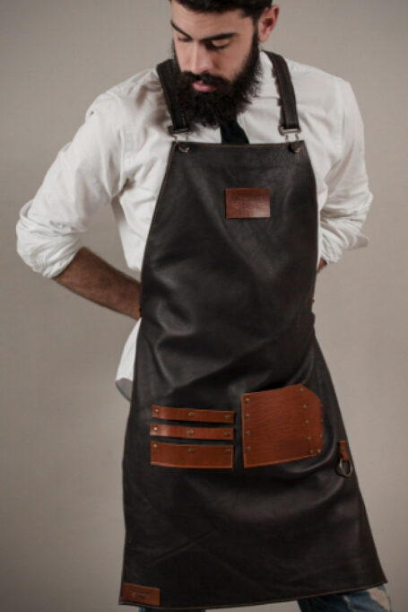 Black leather apron with cross-back straps, pocket, and metal bronze details. Handmade with care. Add a custom logo for a personal touch. Easy to clean and stylish for any workplace.