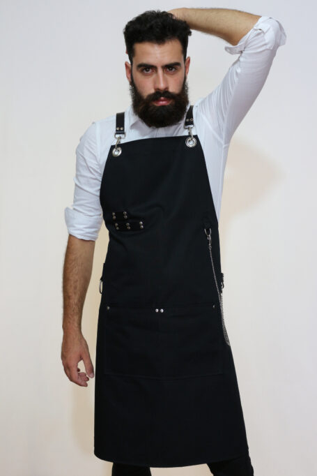 Elegant black polycotton apron with cross-back leather straps, central pocket, and metal accents. Versatile and comfortable for professionals like baristas, chefs, and bartenders. Thoughtful design with divided waist pocket for functionality.