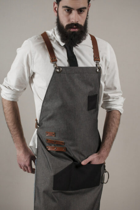 Stylish gray polycotton apron with cross-back straps, leather accents, and black pockets. Ideal for professionals and perfect as a thoughtful gift. Easy to care for and comes in a reusable pouch.