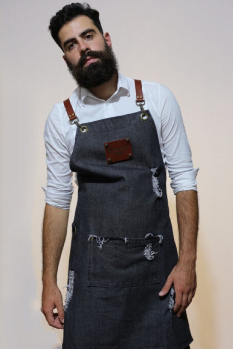 Stylish stonewashed black denim apron with cross-back leather straps, central pocket, and metal bronze details. Versatile and comfortable for professionals like baristas, chefs, and bartenders. Thoughtfully designed for various activities.
