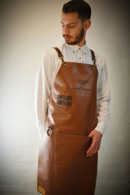 Leather apron in light brown color with adjustable cross-back straps, functional pockets, and unique metal details. Option to add your own custom logo.