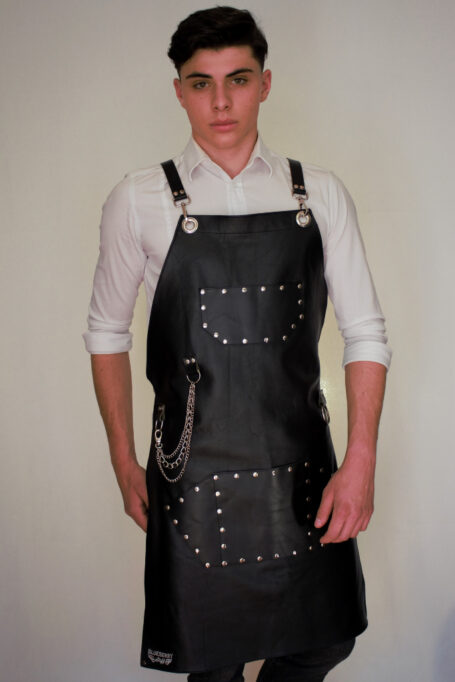 Handmade black leather apron with cross back straps and pockets. Metal chains and rock details. Adjustable Italian cow leather cross back straps in black color. Front view of apron with chest and waist pockets. Unique metal hardware and towel ring on leather apron. Available in sizes for both kids and adults.