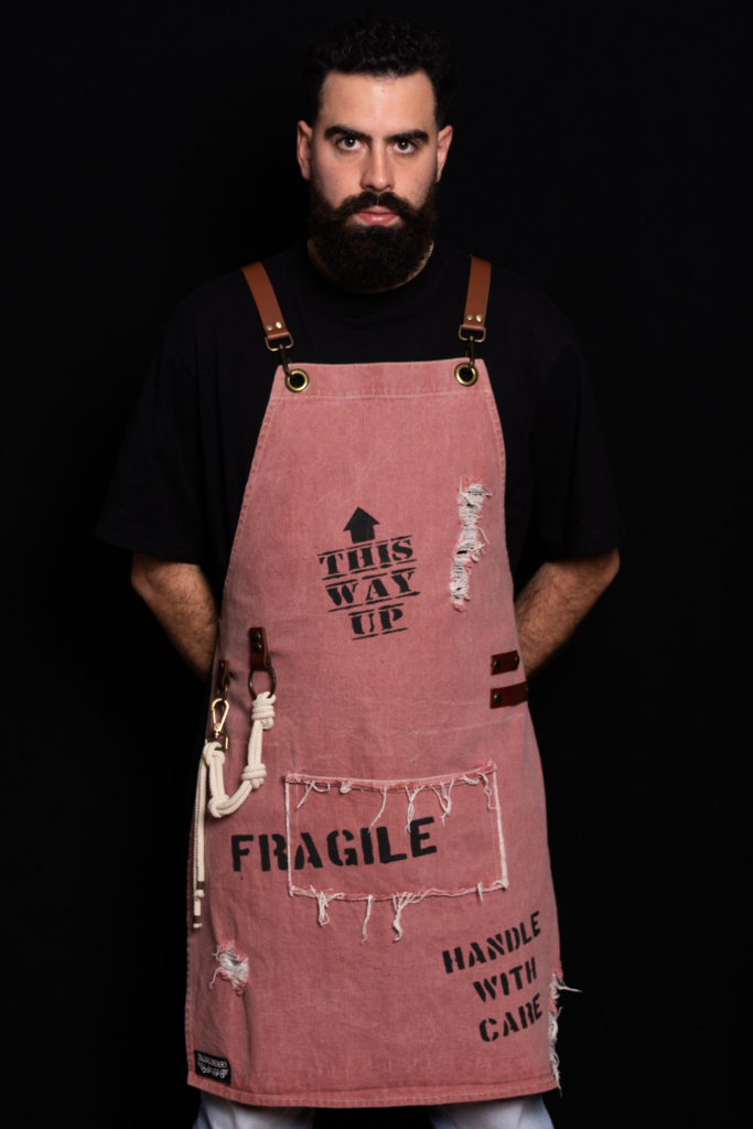 Stylish light maroon denim apron with hand-painted labels, ripped details, and adjustable straps. Features a functional pocket, leather pen holders, and a decorative cotton beige rope. Designed for professionals like baristas, chefs, and more. Available for both kids and parents.
