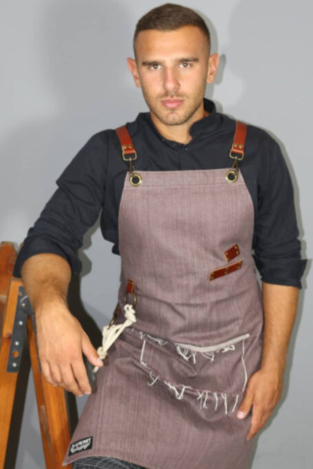 Stylish brownish-purple denim apron with ripped details, cross-back straps, and a convenient pocket. Ideal for professionals like baristas, chefs, and more. Features adjustable leather straps, decorative cotton rope, and easy care instructions. Perfect for kitchen and garden activities.
