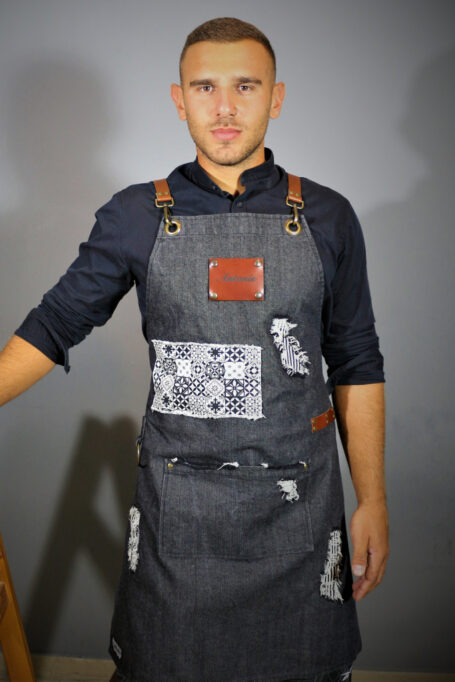 Stylish stonewashed gray denim apron with cross back leather straps, central pocket, and retro tile pattern in black and white. Versatile and comfortable for professionals like baristas, chefs, and bartenders. Available in sizes for both kids and adults.