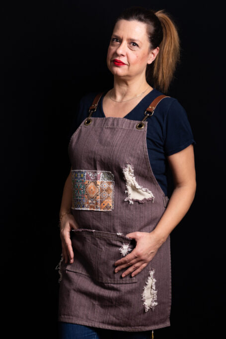 Rusty-brown denim apron with cross-back leather straps, metal bronze accents, and colorful retro tile pattern for a vintage touch. Functional central pocket for tools. Stylish and comfortable attire for professionals like baristas, chefs, and bartenders.