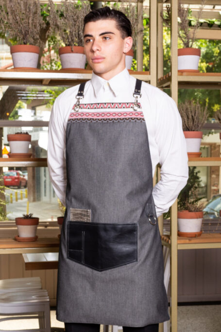 Stylish gray polycotton apron with cross-back leather straps, central black artificial leather pocket, and decorative boho pattern fabric at the chest. Creative and comfortable all-day wear for professionals like baristas, chefs, and bartenders.