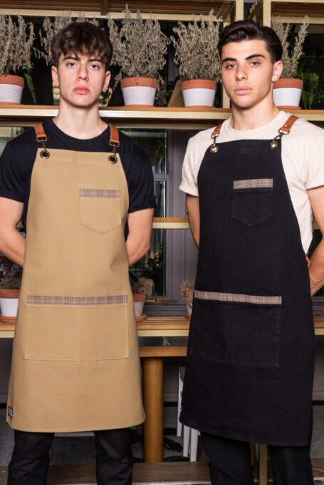 Durable heavy duty apron with cross back leather straps and pockets in black and beige. Ideal for baristas, chefs, and various professionals. Customizable with leather laser logo or embroidered logo options. Easy to clean and makes a thoughtful gift.