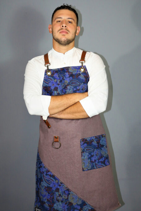Rusty-brown denim apron with cross-back leather straps, metal bronze accents, and blue flower pattern details. Functional pocket in matching fabric for holding tools. Stylish and comfortable for professionals like baristas, chefs, and bartenders.