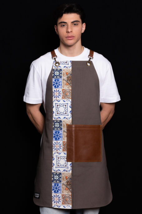 Cotton apron in elephant gray with retro tile pattern, leather pocket, and cross-back straps. Adjustable genuine Italian cow leather straps in light brown. Perfect gift, customizable with your logo. Easy to clean and comfortable for all-day wear.