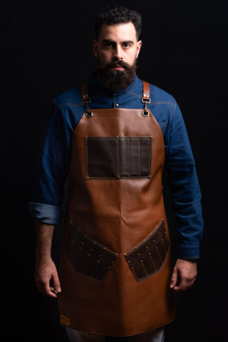 Premium artificial leather apron with pockets, featuring adjustable Italian cow leather cross-back straps and metal bronze details. Ideal for barbers, chefs, and craftsmen. Customizable with laser-engraved logo on chest area.