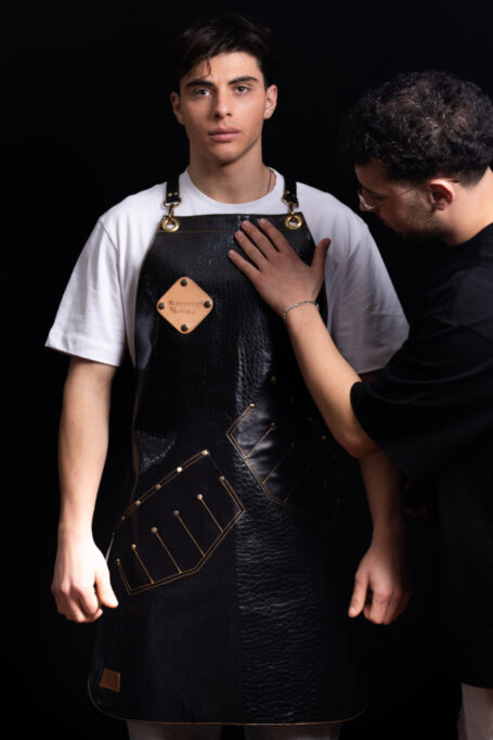 Black leather apron with cross-back straps, asymmetrical pockets, and metal bronze accents. Ideal for barbers, craft makers, and BBQ enthusiasts. Customizable with your logo for a personalized touch.