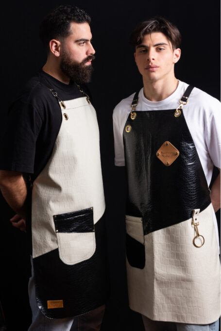Stylish black and white leather apron with cross-back straps, pocket, and towel ring. Perfect for bartenders, barbers, and craftsmen. Customize with your logo for a unique touch. Order now for fast and tracked worldwide shipping.