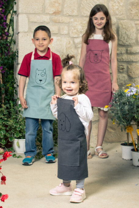 Durable cotton kids’ aprons in different colors, with tied neck straps, pocket and hand painted cute animal faces on the chest area. Playful attire.