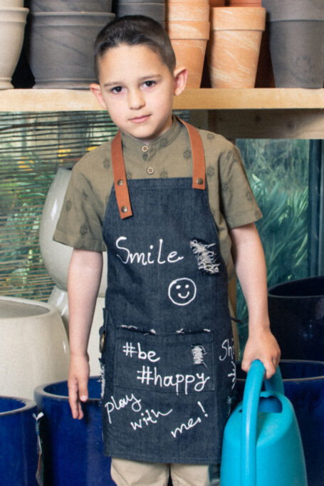 Exclusive stonewashed denim kids’ apron in gray color, with adjustable straps, pocket and hand-painted drawings and quotes. Trendy and street style vibes.