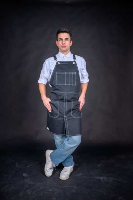 A stylish man stands against a black backdrop, donned in a white shirt and a custom black apron. Intricate white threadwork adds flair to the medium-weight cotton fabric, blending practicality and elegance seamlessly.