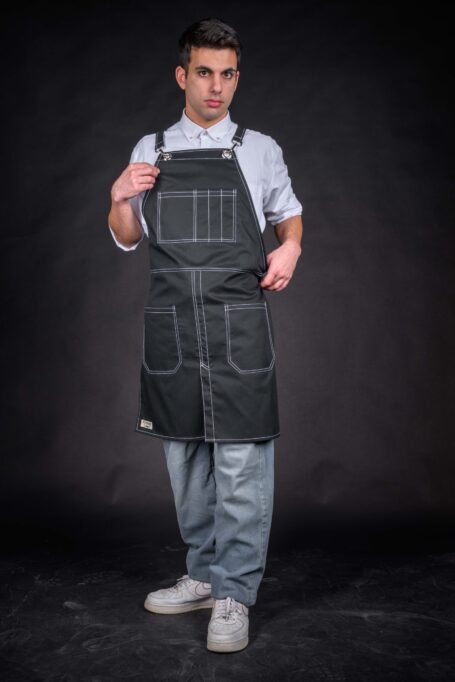 In this image, a man exudes sophistication in a white shirt and a custom black apron against a black background. The intricate white threadwork on medium-weight cotton fabric adds a personalized touch, marrying style with practicality.