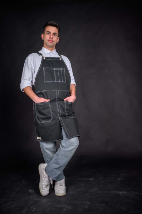 Against a black canvas, a man wears a white shirt paired with a custom black apron. Beautiful white threads adorn the medium-weight cotton fabric, creating a visually striking ensemble for culinary or artisanal pursuits.
