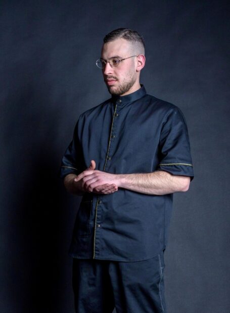 Hair stylist wearing his black workwear jacket with metal bronze stud buttons and gold brocade on front placket, pocket and around the sleeves.