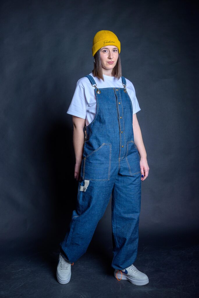 Overall baggy street style unisex pants. Woman wearing a blue denim dungaree with metal bronze stud buttons.