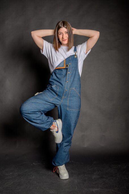 Happy woman wearing a comfortable salopette, with external decorative orange stitches and two front pockets.