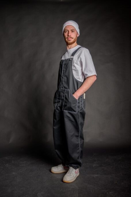 Man wearing streetwear unisex black salopette, which has two front pockets and adjustable straps.